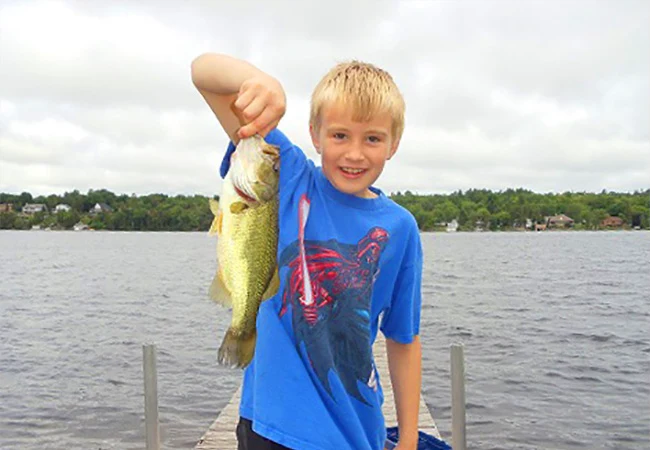 Child holding a Largemouth Bass while standing on a dock by a lake