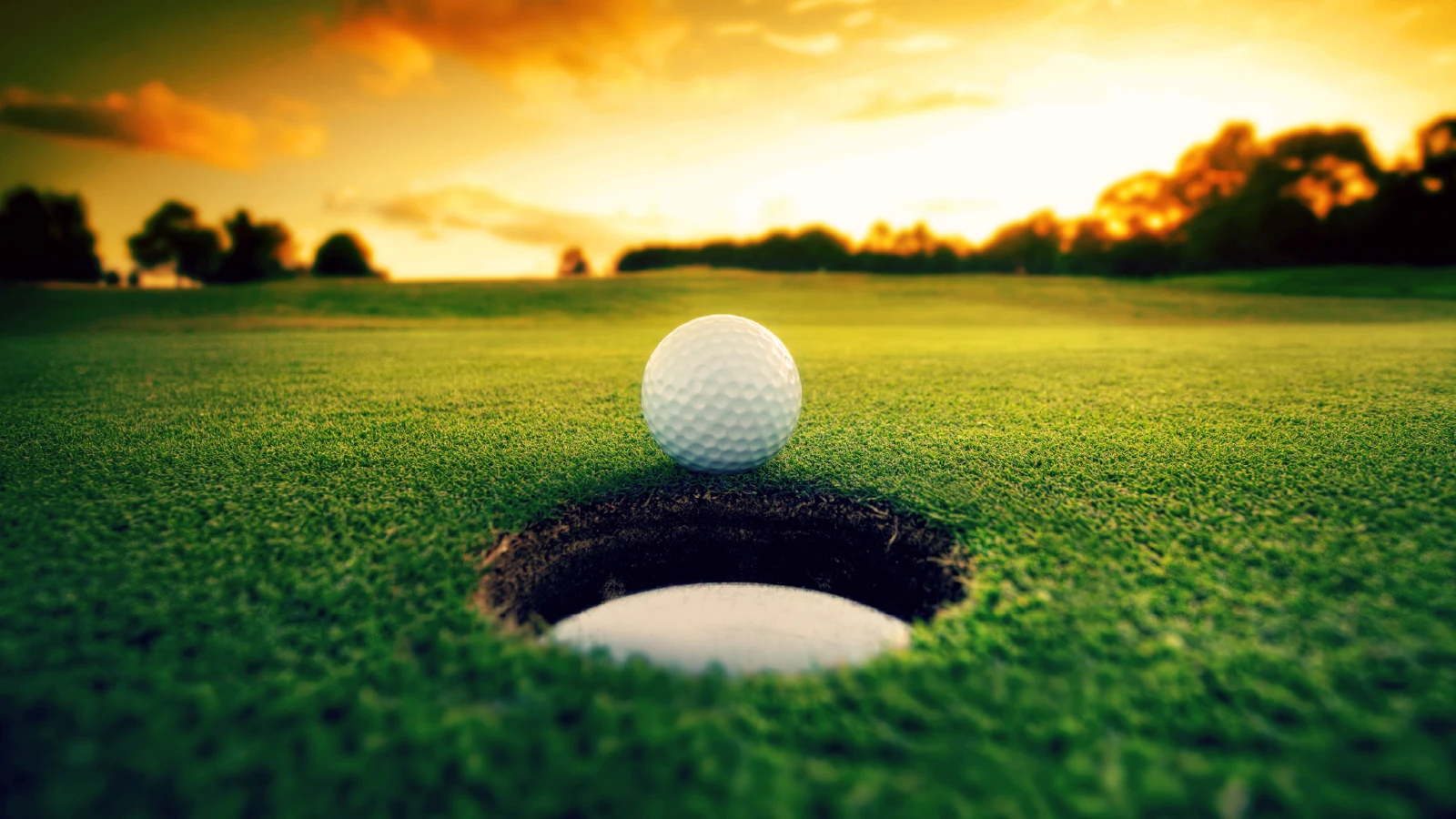 Golf ball positioned on the edge of a hole on a green fairway at sunset, representing golfing near Pigeon Lake in Kawartha Lakes Ontario