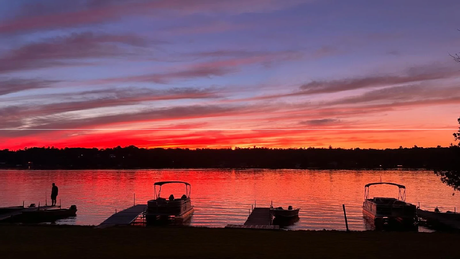 Pigeon Lake sunset with vibrant red and orange sky reflecting on calm water, boats docked along shoreline at Pigeon Lake cottages in Kawartha Lakes Ontario